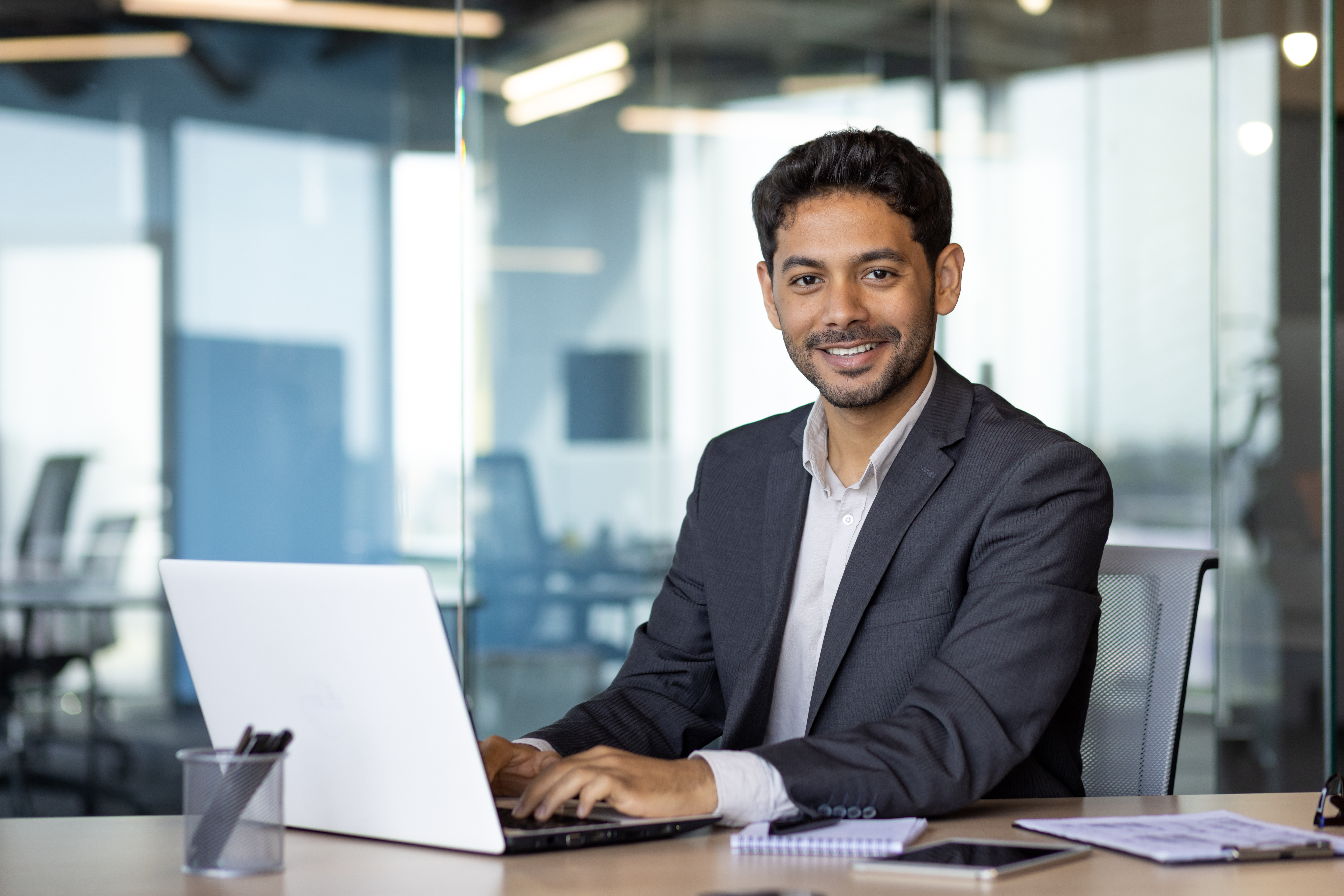 Portrait of young arab businessman, man smiling and looking at camera while sitting inside office, boss in business suit at workplace using laptop